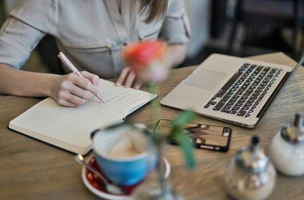 Imagem de uma mulher na frente de um notebook, escrevendo em um caderno para ilustrar artigo sobre escrever bem. Foto: Pexels.