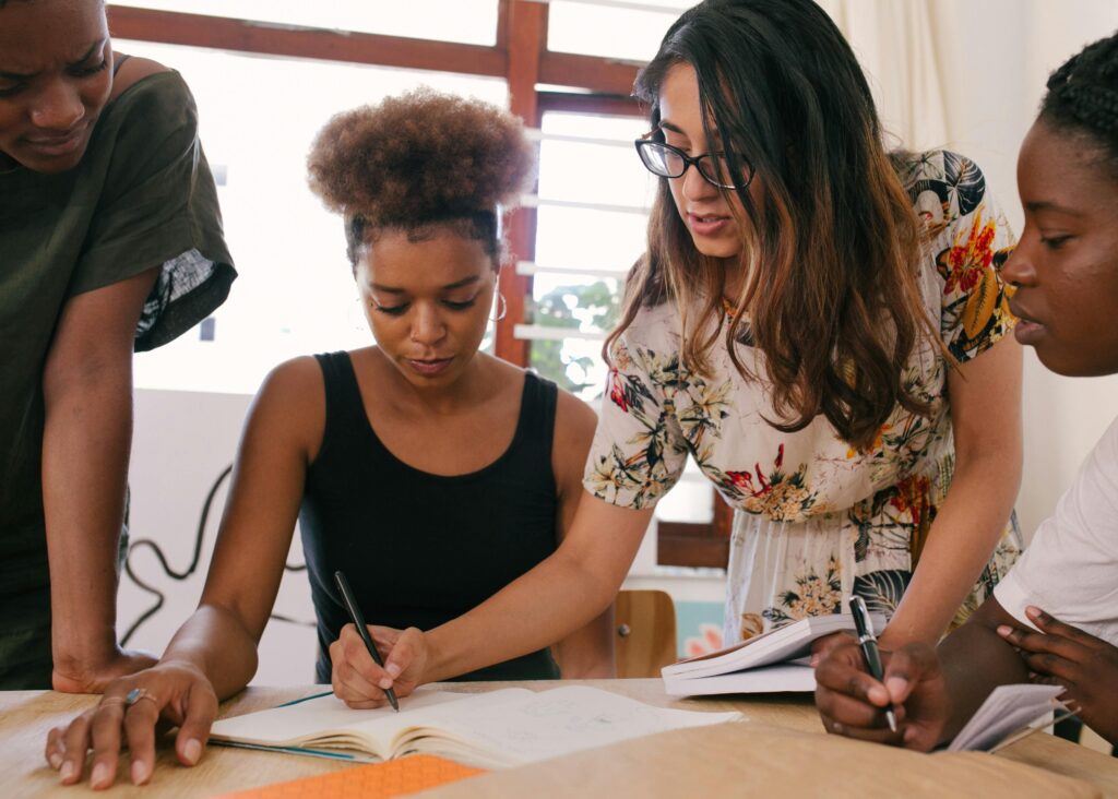 Imagem de quatro pessoas reunidas. Uma delas escreve em um caderno. Foto: Pexels.